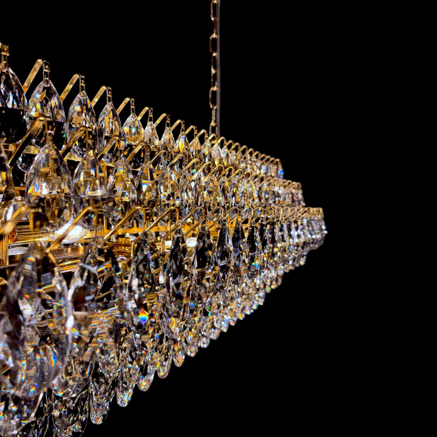 A detailed side perspective view of a linear crystal chandelier with a  gold frame, highlighting the multiple layered tiers of teardrop crystals against a solid black background.
