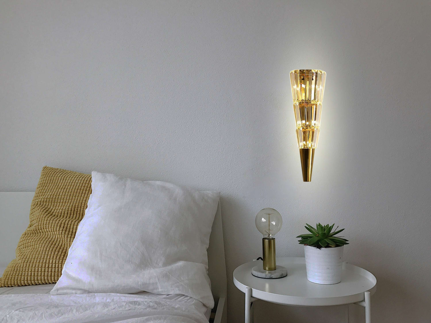 Gold cone-shaped wall lamp on a gray wall above a bed with white bedding and a small table with decorative items.