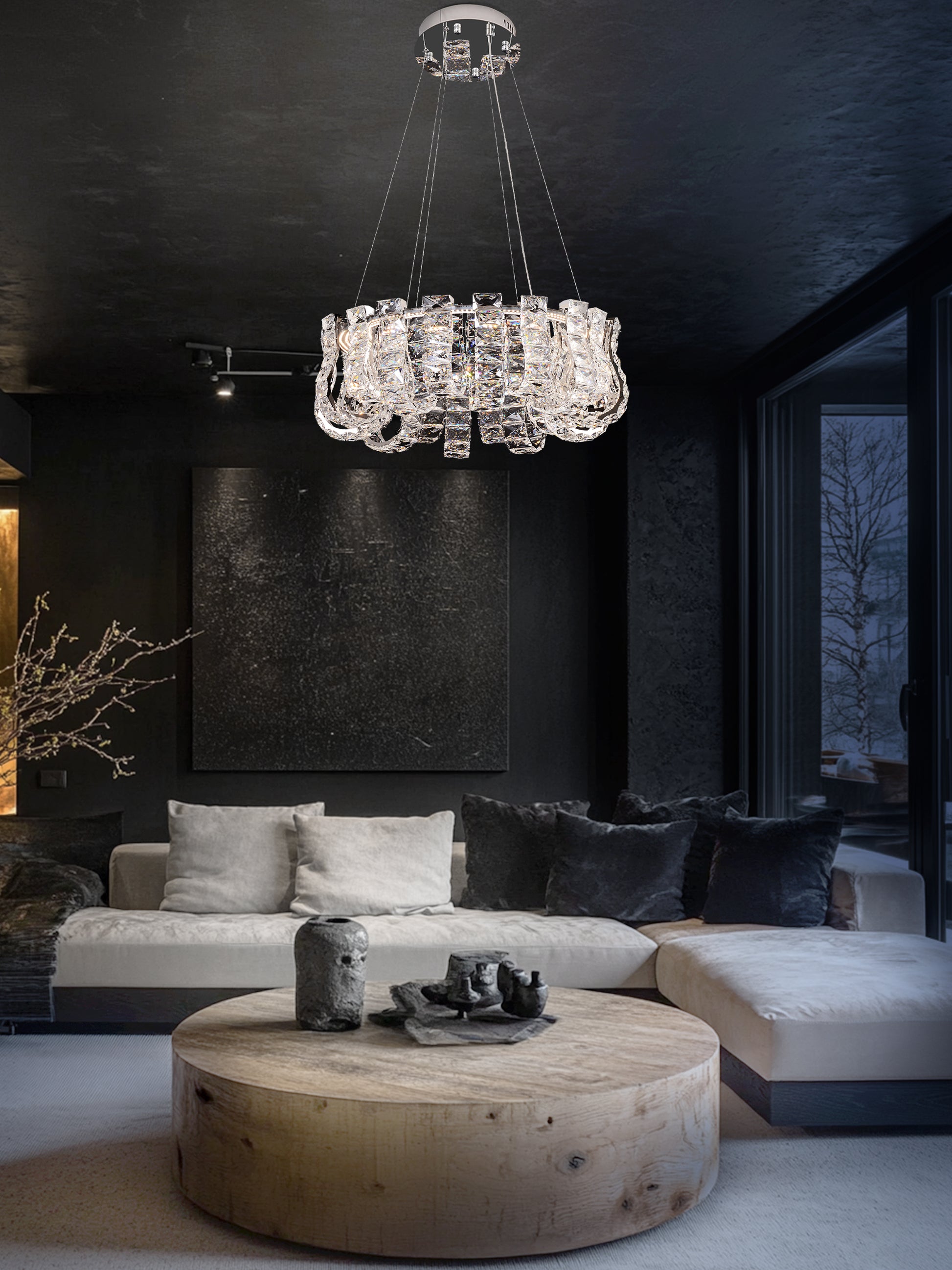 A modern crystal LED chandelier with a chrome finish hanging in a room with dark walls and a round wooden table in the foreground.