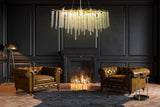 A luxurious gold crystal raindrop chandelier hanging from the ceiling, with a branch-inspired design, displayed in a room with two leather chairs and a fireplace in the background.