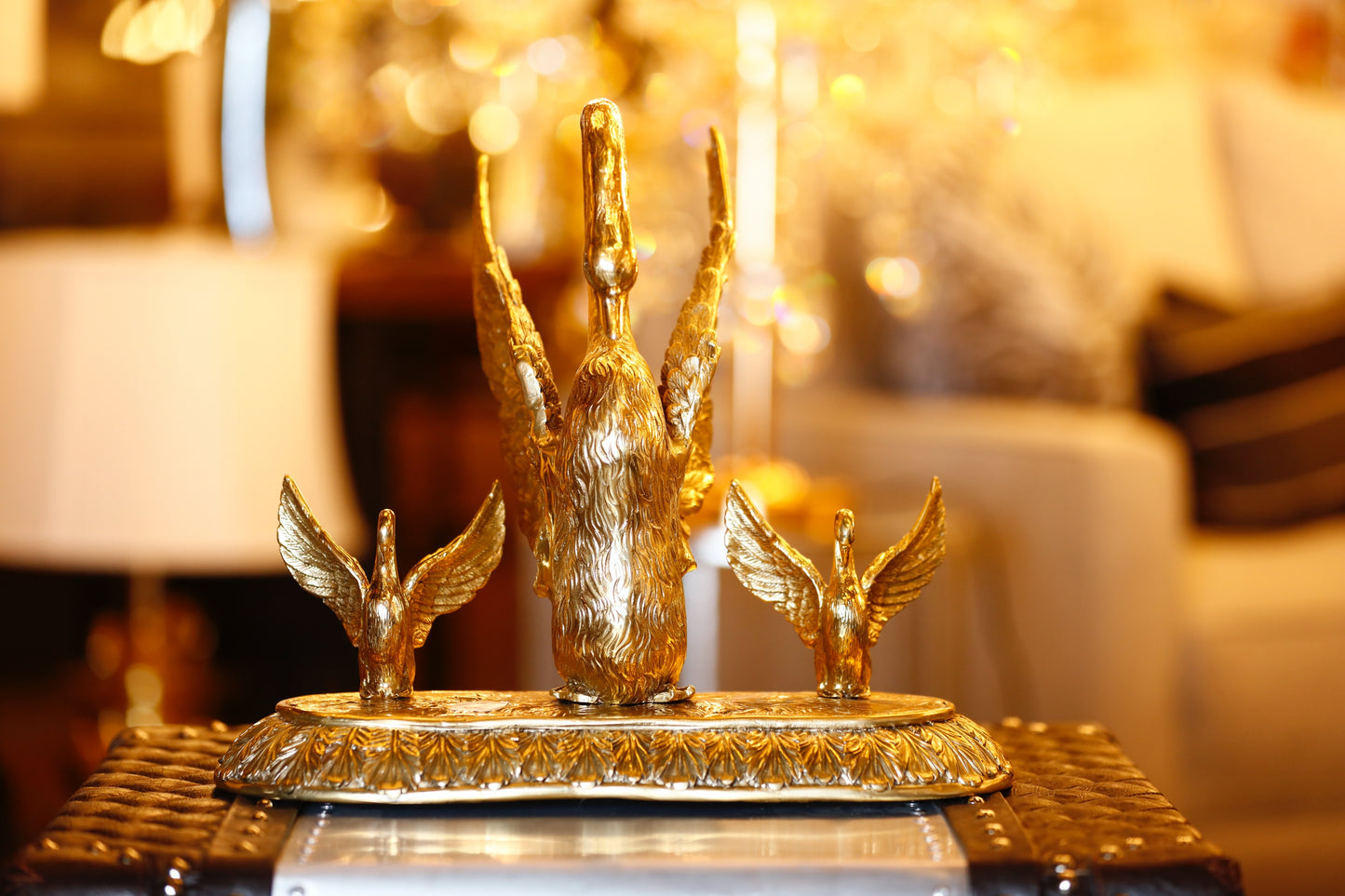 A brass sculpture of a golden swan family with one larger swan and two smaller ones, placed on a circular base, displayed in a brightly lit interior setting.