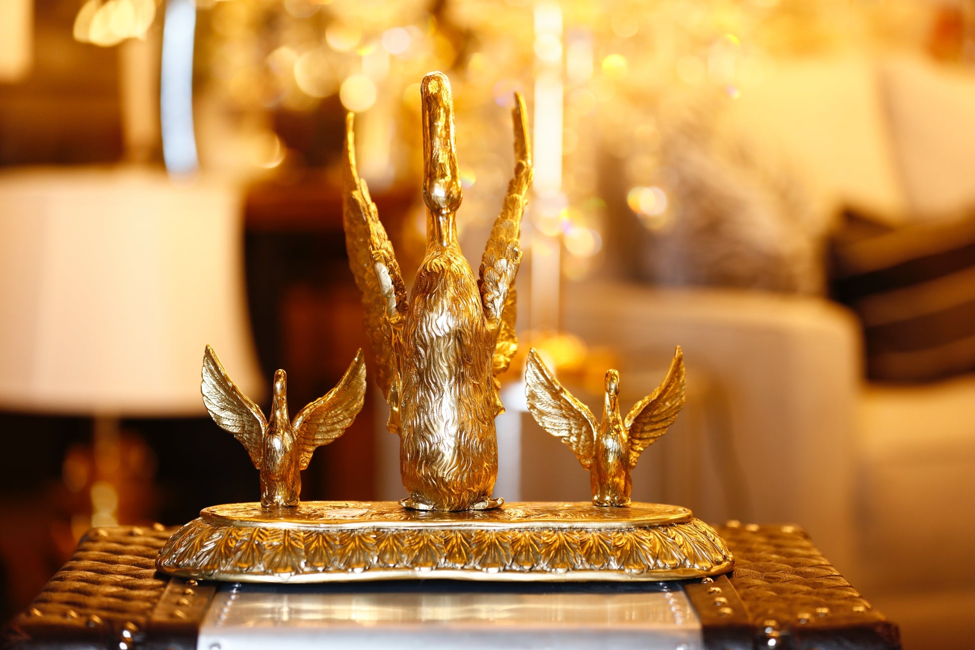 A brass sculpture of a golden swan family with one larger swan and two smaller ones, placed on a circular base, displayed in a brightly lit interior setting.