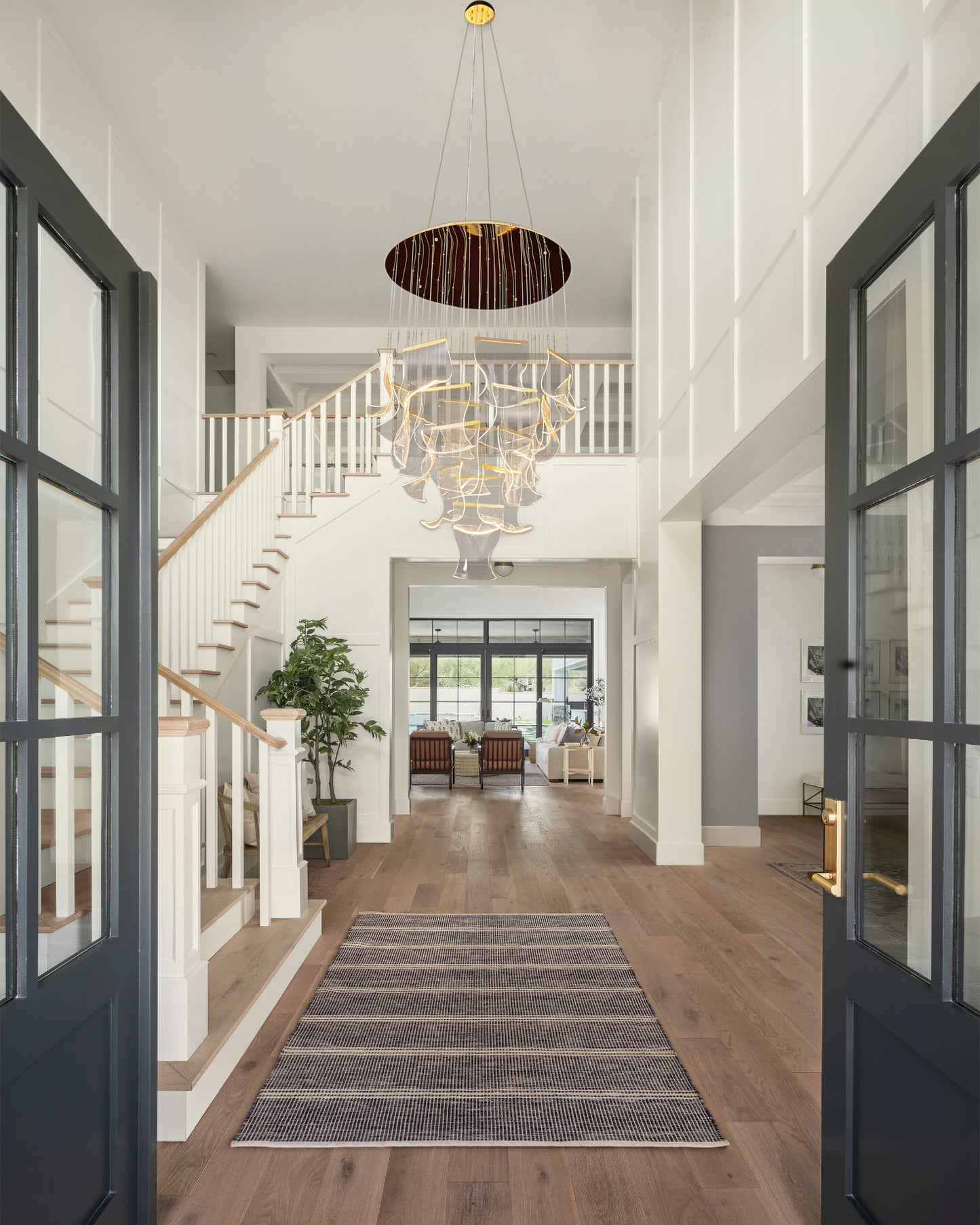 Dimmable gold LED chandelier with adjustable hanging cables lighting up a tall entryway with staircase and high ceiling.