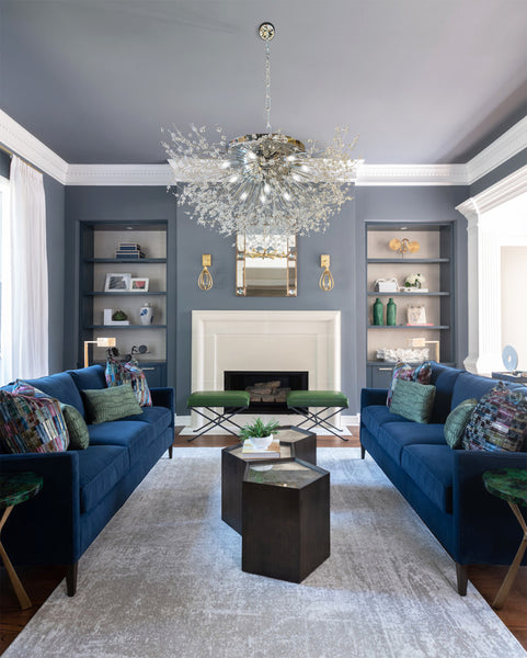A luxurious silver chrome chandelier with a starburst design, featuring nano-cut crystal beads, hanging over a living room setting with two blue sofas and a fireplace in the background.